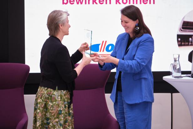 Zwei Frauen stehen sich gegenüber und lächeln, während eine von ihnen der anderen eine gläserne Auszeichnung überreicht.