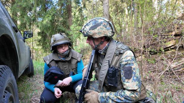 Zwei Personen in Schutzwesten und Helmen knien im Wald neben einem Militärfahrzeug.