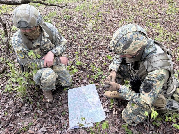 Zwei Soldaten in Tarnuniform knien im Wald und orientieren sich mit einer Karte und einem Kompass.