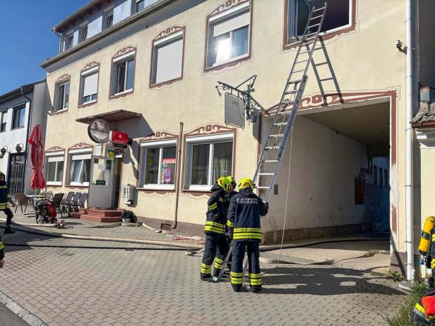 Drei Feuerwehrleute stehen vor einem Gebäude mit Leiter am Fenster, Schläuchen und Ausrüstung auf dem Gehweg.