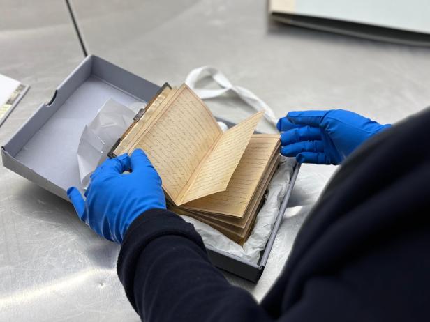 Eine Person mit blauen Handschuhen blättert vorsichtig in einem alten handschriftlichen Buch in einer Schachtel.