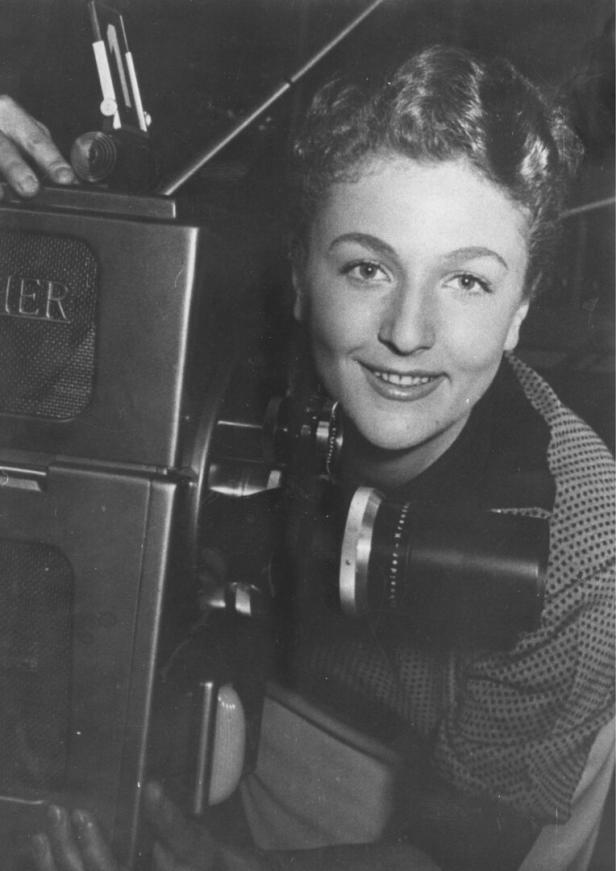 Eine lächelnde Frau steht neben einer alten Fernsehkamera.