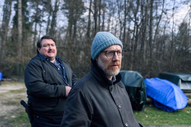 Zwei Männer stehen im Freien vor abgedeckten Gegenständen, einer trägt eine blaue Mütze und Brille, der andere eine dunkle Jacke.