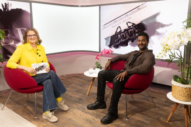 Zwei Personen sitzen in roten Sesseln in einem modernen Studio mit Pflanzen und großen Bildschirmen im Hintergrund.