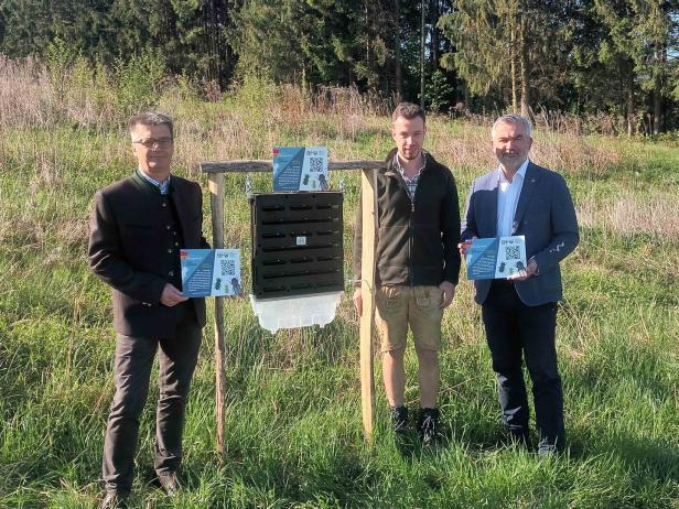 Drei Männer stehen auf einer Wiese neben einer Insektenfalle und halten Infotafeln mit QR-Codes in den Händen.
