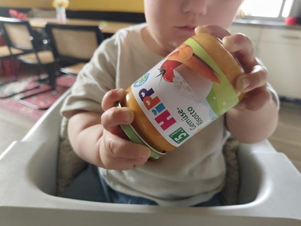 Ein Kleinkind hält ein Glas mit Hipp Gemüse Risotto Babynahrung in der Hand.