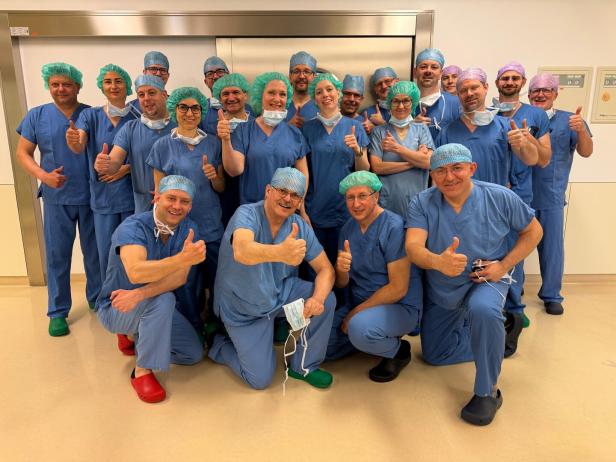 Eine Gruppe von medizinischem Personal in blauer OP-Kleidung und Hauben zeigt lächelnd den Daumen nach oben.