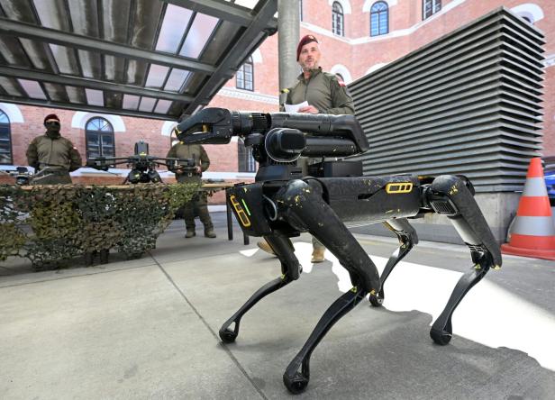 Ein vierbeiniger Roboter steht vor Soldaten in Uniform, im Hintergrund militärische Ausrüstung und ein Tarnnetz.