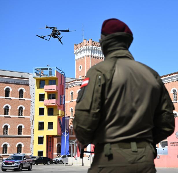Ein Soldat beobachtet eine fliegende Drohne vor einem historischen Gebäude mit buntem Anbau und mehreren geparkten Autos.
