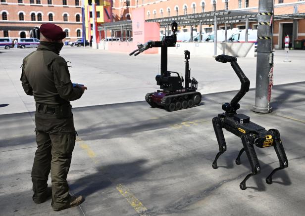 Ein Polizist steuert zwei verschiedene Einsatzroboter auf einem Hof, einer mit Kettenantrieb und Greifarm, der andere mit vier Beinen.