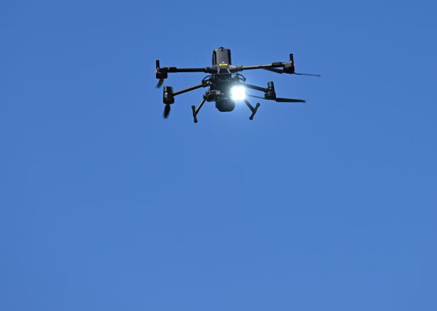 Eine Drohne mit eingeschaltetem Licht fliegt vor klarem, blauem Himmel.