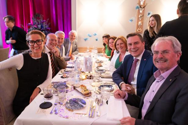 Mehrere elegant gekleidete Menschen sitzen lachend an einer festlich gedeckten Tafel in einem Restaurant.