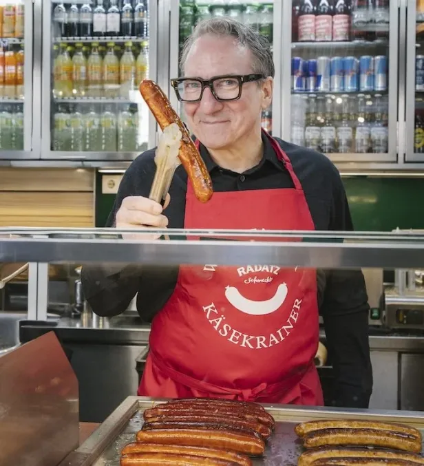 Mann mit roter Schürze hält eine Käsekrainer-Wurst mit einer Zange vor einer Wursttheke.