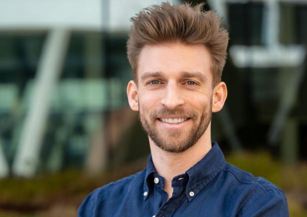 Ein junger Mann mit braunen Haaren und Bart lächelt freundlich und trägt ein dunkelblaues Hemd.