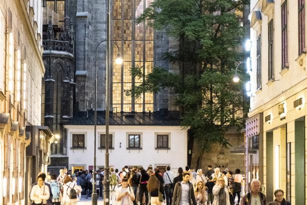 Menschen flanieren am Abend durch eine belebte Gasse in Wien, im Hintergrund die beleuchteten Fenster des Stephansdoms.