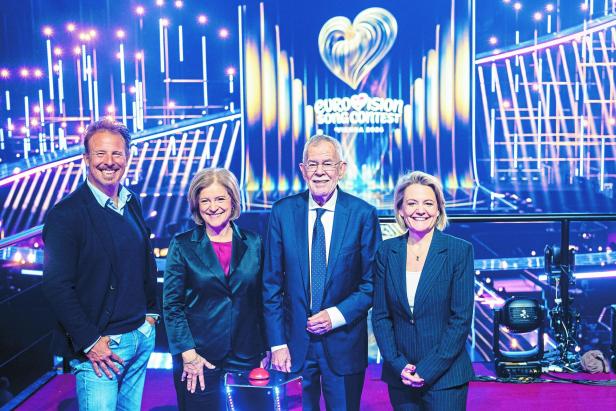 Vier Personen stehen lächelnd vor einer bunten Bühne mit Eurovision Song Contest Logo und Lichteffekten.