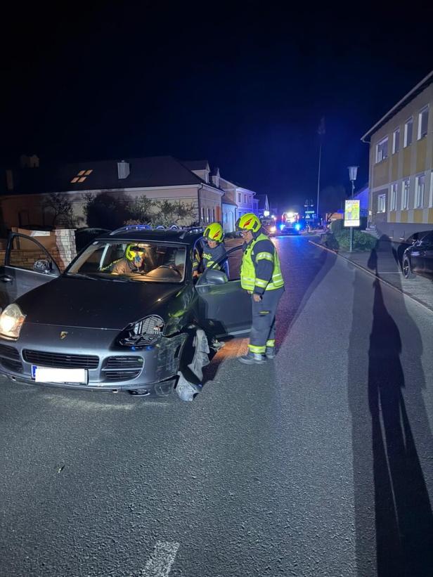 Zwei Feuerwehrleute stehen nachts neben einem beschädigten Auto auf einer Straße, Blaulichter sind im Hintergrund zu sehen.