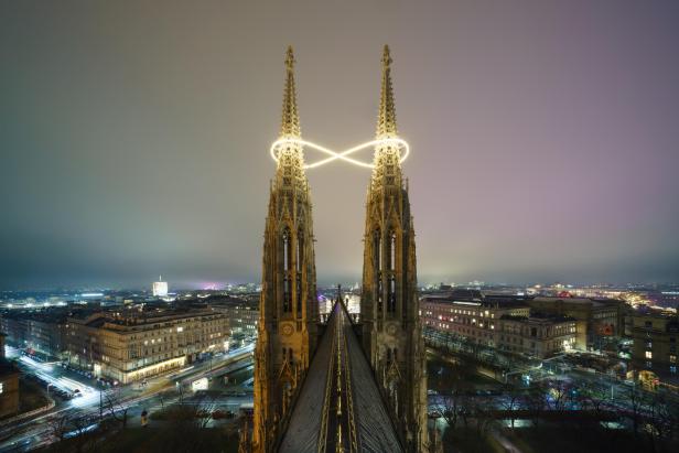 Die beiden Türme der Votivkirche in Wien sind nachts beleuchtet und mit einer leuchtenden Kunstinstallation verbunden, während die Stadt im Hintergrund erstrahlt.
