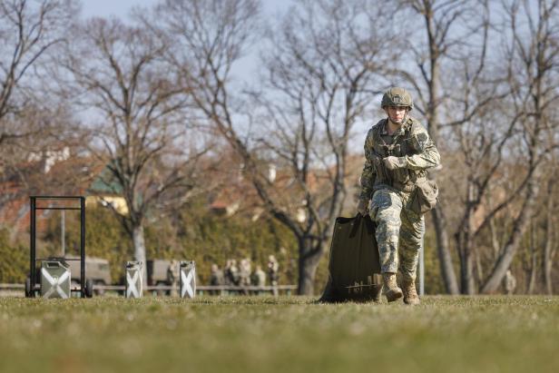 Soldat in Tarnuniform trägt Ausrüstung über eine Wiese, im Hintergrund weitere Soldaten.
