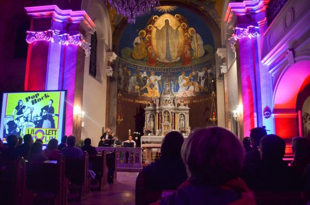 Konzertbesucher erleben in einer bunt beleuchteten Kirche eine Pop-Rock-Show mit Band und Projektion, während der Altarraum festlich illuminiert ist.