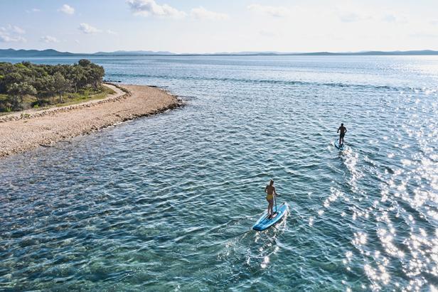 Zwei Personen paddeln auf SUP-Boards über das klare Meer entlang einer bewaldeten Küste.