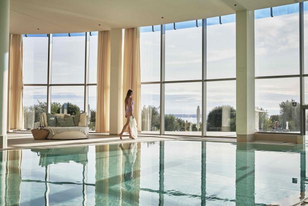 Frau im Badeanzug spaziert am Innenpool mit Blick auf Garten und Meer durch große Fenster.