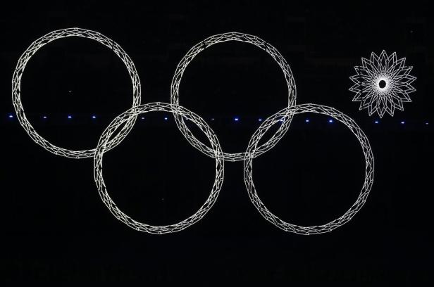 Die olympischen Ringe und eine stilisierte Blume leuchten vor schwarzem Hintergrund.
