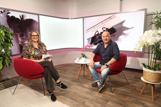Zwei Personen sitzen in roten Sesseln in einem modern eingerichteten Studio mit Pflanzen und großen Bildschirmen im Hintergrund.