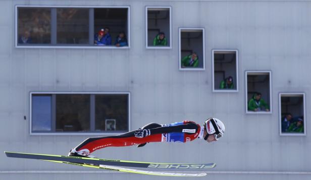 Ein Skispringer in der Luft vor einem Gebäude mit Zuschauern in den Fenstern.