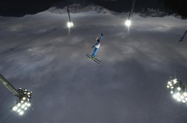 Ein Skifahrer springt nachts vor dem Hintergrund von Bergen und Flutlichtmasten.
