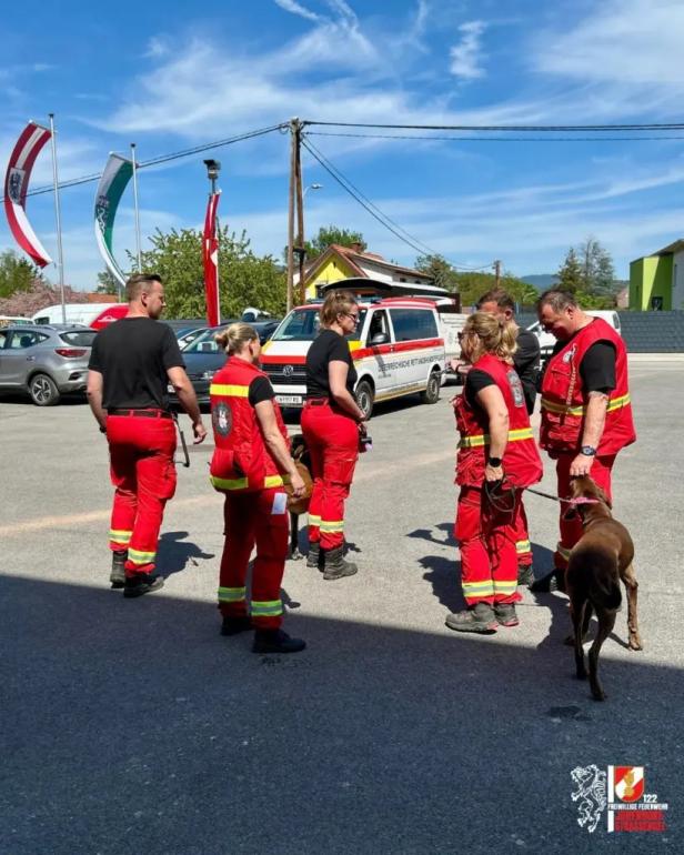 Mehrere Rettungskräfte in roten Uniformen mit Suchhunden auf einem Parkplatz bei Sonnenschein.