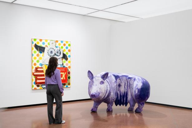 Ausstellungsansicht "Animalia", eine Frau in einem violetten Pullover betrachtet die Skulptur von einem Schwein, das dieselbe Farbe wie ihr Pullover hat, sie steht vor einem knallbunten Bild