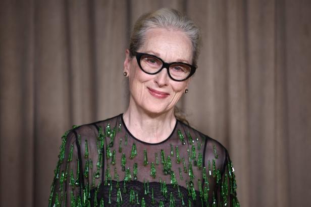 Meryl Streep lächelt mit Brille und grün verziertem Oberteil vor beigem Hintergrund.
