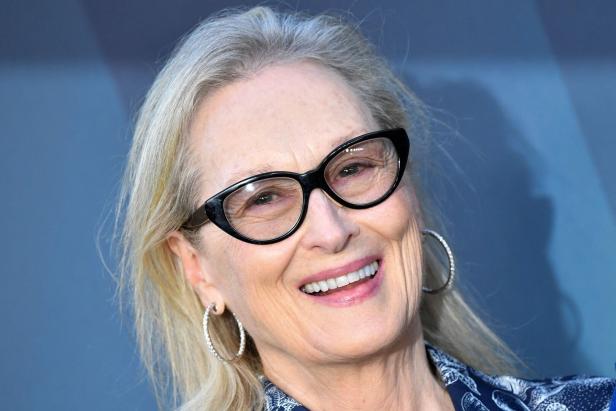 Meryl Streep lächelt mit Brille und Ohrringen vor blauem Hintergrund.