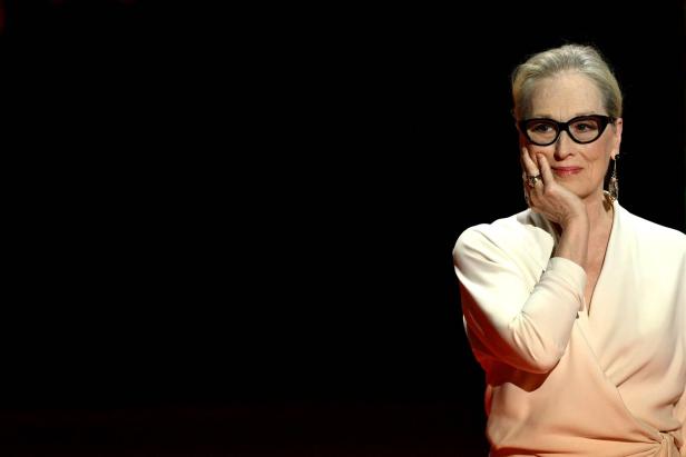 Meryl Streep mit Brille und hellem Oberteil posiert vor dunklem Hintergrund.