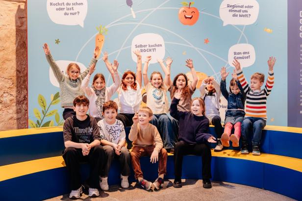 Gruppe fröhlicher Kinder sitzt und jubelt vor einer bunten Wand mit Gemüse-Illustrationen.