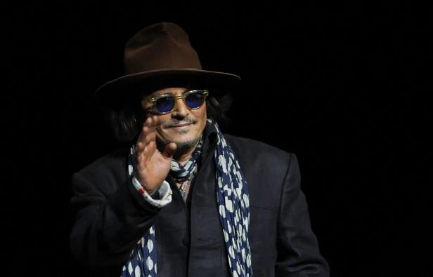Johnny Depp mit Hut, Schal und Sonnenbrille winkt vor dunklem Hintergrund.