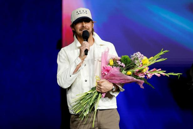 Ryan Gosling mit Kappe hält ein Mikrofon und einen Blumenstrauß vor einem blauen Hintergrund.