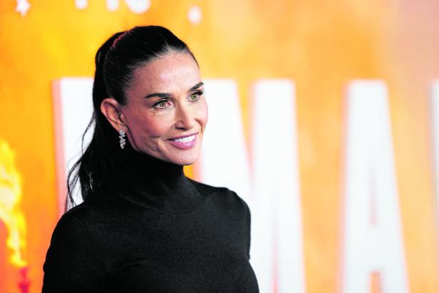 Demi Moore mit hochgesteckten, dunklen Haaren und schwarzem Rollkragenpullover lächelt.