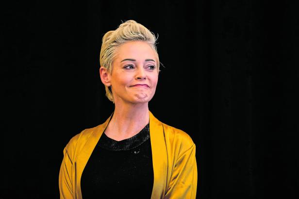 Rose McGowan mit kurzen blonden Haaren und goldener Jacke blickt lächelnd zur Seite.