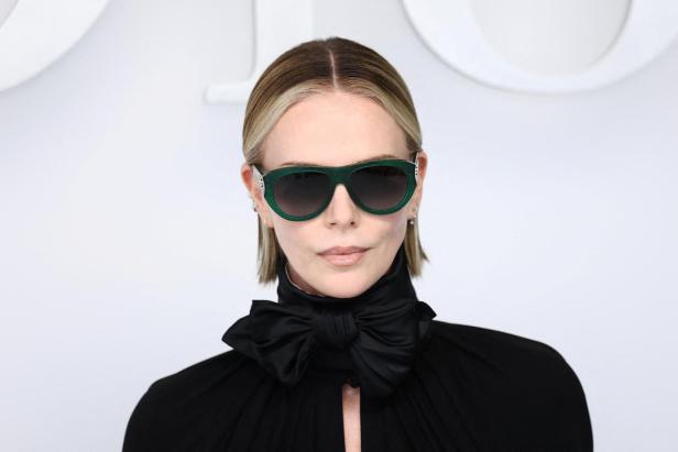 Charlize Theron mit blonden Haaren trägt eine grüne Sonnenbrille und eine schwarze Bluse mit Schleife.