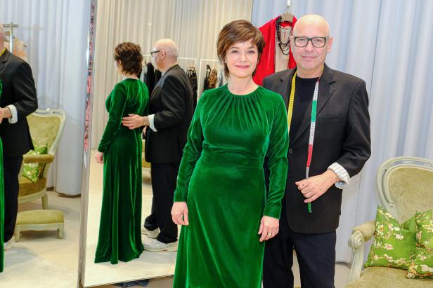 Eine Frau im grünen Samtkleid steht neben einem Mann mit Maßband um den Hals in einem eleganten Ankleidezimmer.