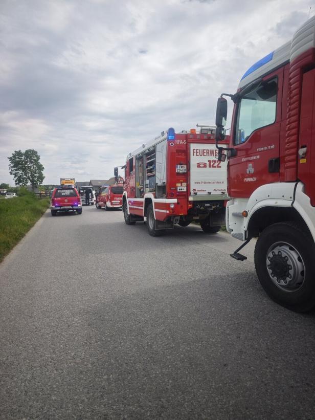 Mehrere Feuerwehrfahrzeuge stehen auf einer Landstraße, Einsatzkräfte sind im Hintergrund im Einsatz.