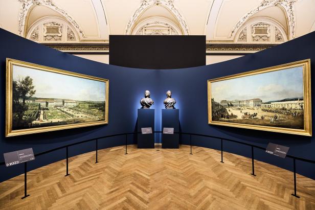 Zwei großformatige Veduten von Schloss Schönbrunn und belebtem Vorplatz flankieren Büsten von Maria Theresia und Franz I. Stephan in der Ausstellung „Canaletto & Bellotto“ im Kunsthistorischen Museum.
