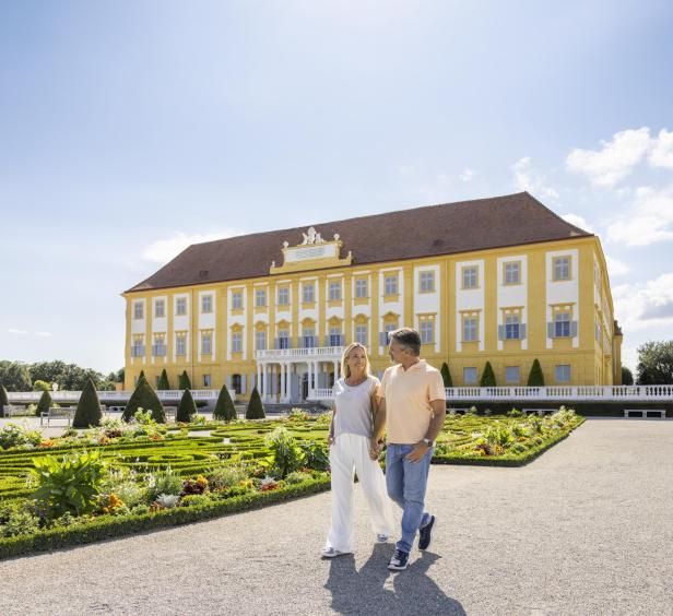 Ein Paar spaziert durch den barocken Garten vor dem gelben Schloss Hof bei Sonnenschein.
