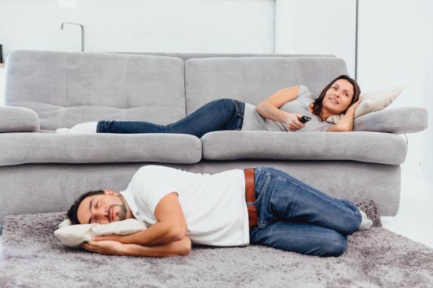 Eine Frau entspannt auf einem grauen Sofa mit Fernbedienung, während ein Mann davor auf dem Teppich liegt und schläft.