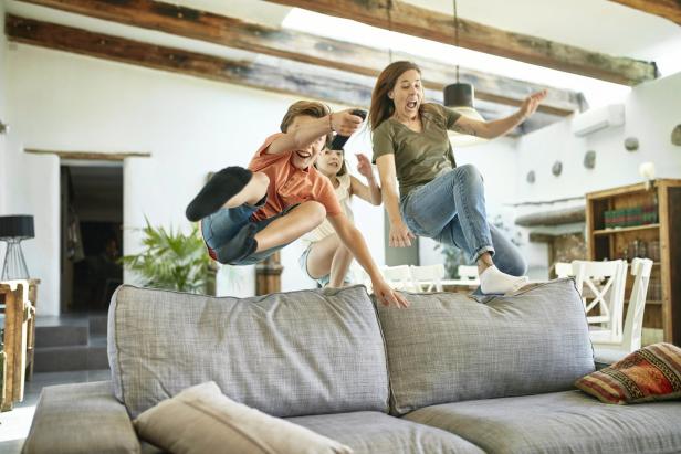 Eine Frau und zwei Kinder springen lachend auf ein großes, graues Sofa im Wohnzimmer und zeigen damit die Alltagstauglichkeit und Robustheit der Couch.