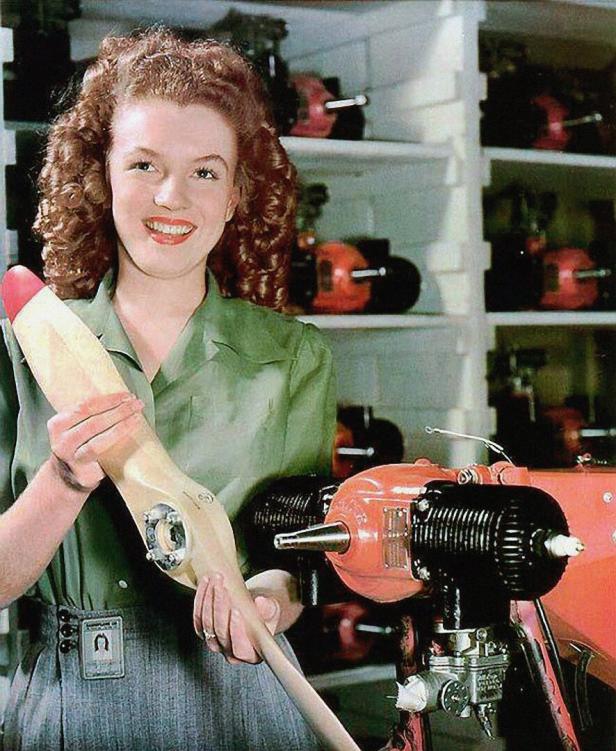 Eine lächelnde Frau mit lockigem Haar hält einen Propeller vor einem Regal mit Motoren.