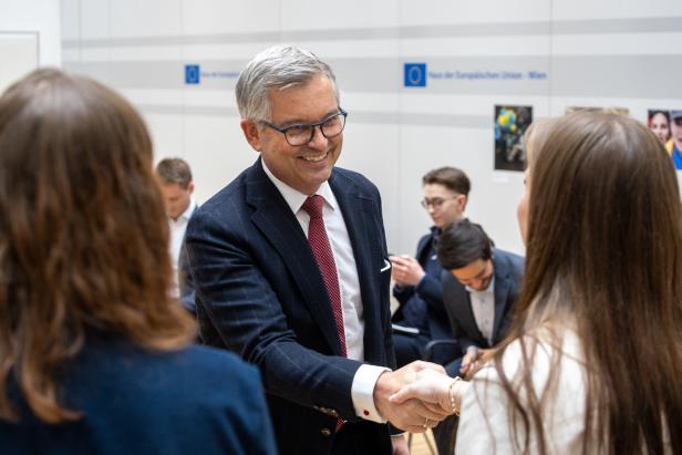 Magnus Brunner (EU Kommissar) im Anzug gibt einer jungen Frau die Hand und lächelt, im Hintergrund sitzen weitere Personen.