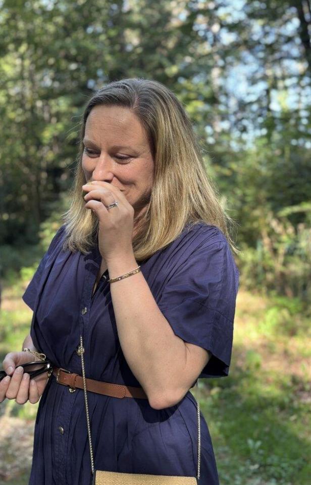 Eine Frau mit blonden Haaren steht lachend im Sonnenschein vor einem grünen, bewaldeten Hintergrund.